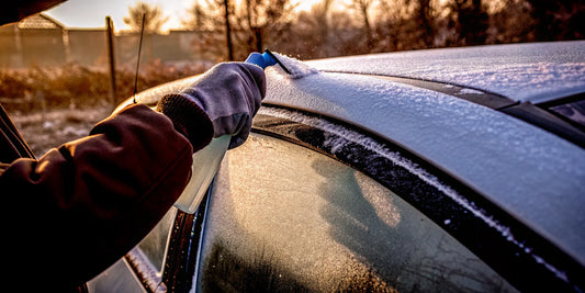 A gloved hand using a scraper to de-ice a car's frozen wiper blades.