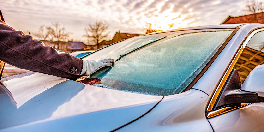A gloved hand scraping a frozen car windshield, a task you can avoid by stopping it from freezing.