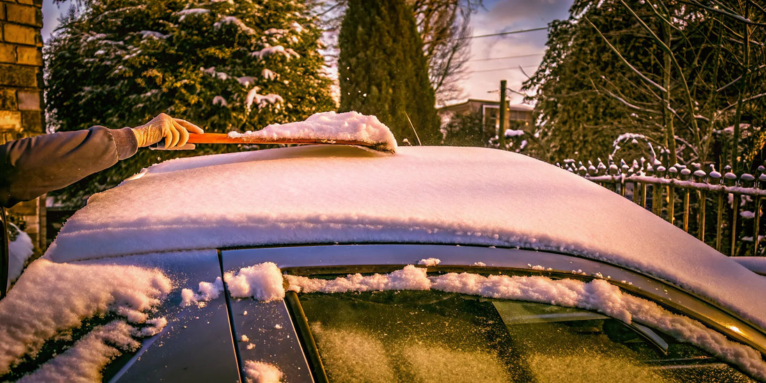 Driver sweeping snow off a car roof with a wide orange scraper in a snowy driveway
