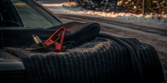Winter car emergency kit items including an ice scraper, blanket, flashlight, and jumper cables arranged on a snowy vehicle hood