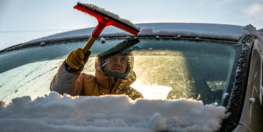 Driver clearing snow from a car windshield with a large red super-sized snow scraper on a winter morning